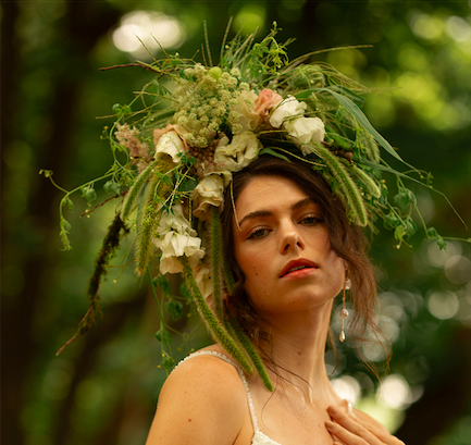 Amidst a forest background, a young woman with brown hair wears a fantastical floral headpiece created by District 2 Floral Studio incorporating locally grown flowers for a mid-August fashion editorial shoot for Nebraska Wedding Day magazine. 