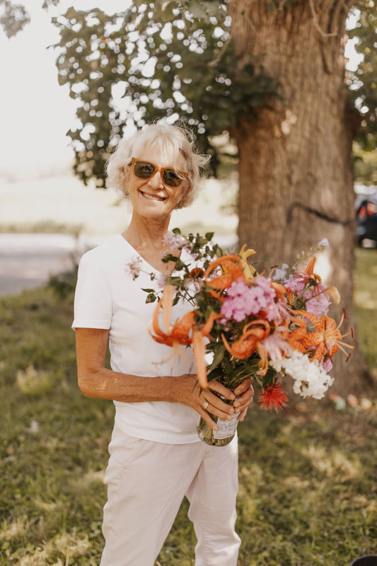 Adult smiling wearing sunglasses under a tree holding a flower arrangement they created during a community flower workshop with District 2 Floral Studio.