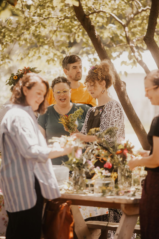 Adults smiling around a table surrounded by trees while they each create something with flowers during a community flower workshop with District 2 Floral Studio.