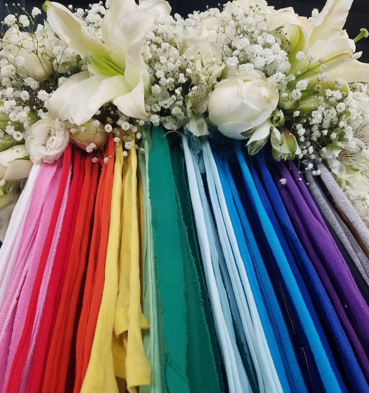 Up close of white lilies and baby's breath form a cloud shape with fabric strips in a rainbow spectrum of colors falling below the the flowers. Created by District 2 Floral Studio.