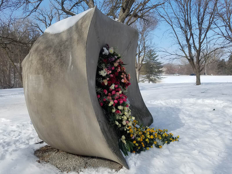 Side view of a park sculpture, Sounding  Stones, with pink, white and yellow flowers cascading out of its center. The flowers are bright against the white snow. Installation by District 2 Floral Studio.