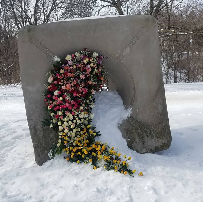A park sculpture, Sounding  Stones, has pink, white and yellow flowers cascading out of its center. The flowers are bright against the white snow. Installation by District 2 Floral Studio.