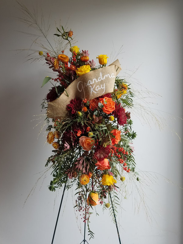 Foam-free floral funeral spray on an easel with warm colors and grasses with a banner across it that reads, "Grandma Kay". Created by District 2 Floral Studio.