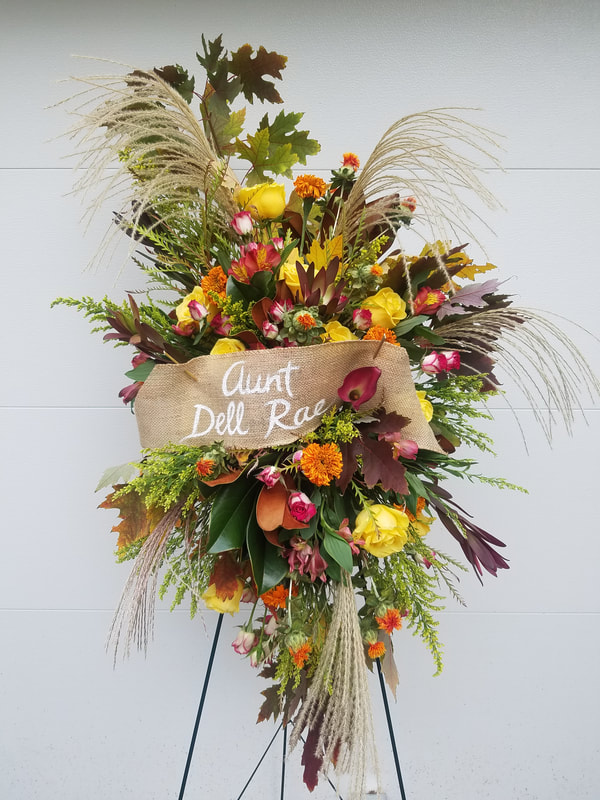 Foam-free floral funeral spray on an easel with warm colors and grasses with a banner across it that reads, "Aunt Dell Rae". Created by District 2 Floral Studio.