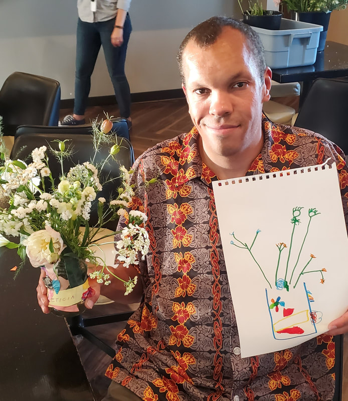 Adult participant holds a flower arrangement and drawing of flowers created during a District 2 Floral Studio flower design and observational drawing workshop.