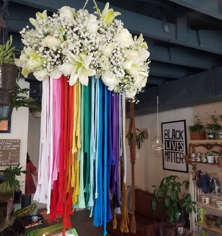 White lilies and baby's breath form a cloud shape with fabric strips in a rainbow spectrum of colors falling below the the flowers. Created by District 2 Floral Studio.