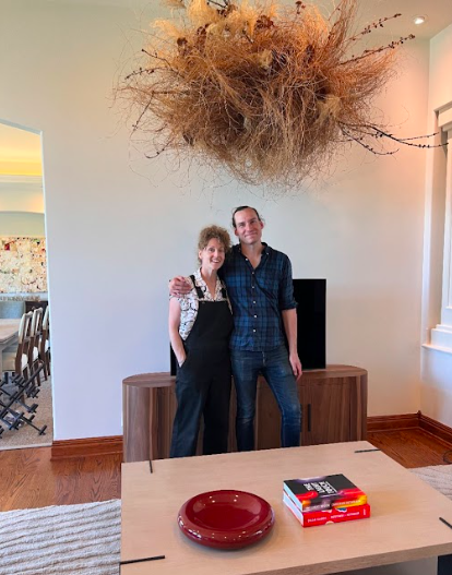 Branches and locally grown Nebraska dried grasses form a cloud created by District 2 Floral Studio that is suspended from the ceiling above a TV in a spacious living room. Two humans stand under it.