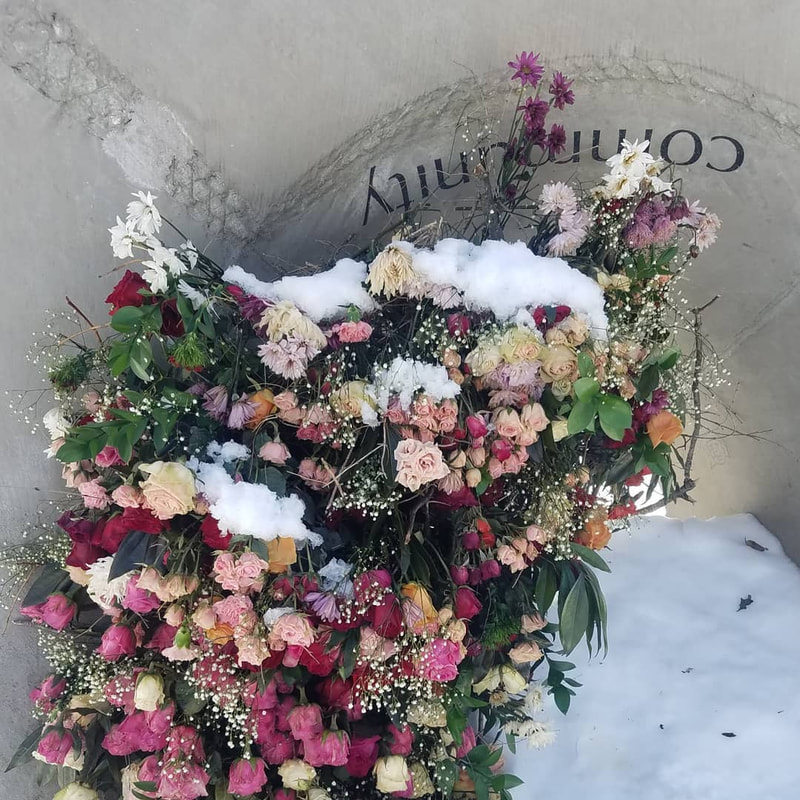 A park sculpture, Sounding  Stones, has pink, white and yellow flowers cascading out of its center. The word "community" is engraved in the sculpture. The flowers are bright against the white snow. Installation by District 2 Floral Studio.